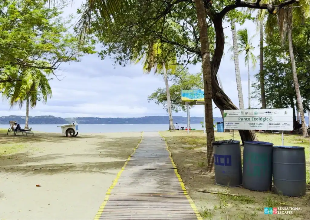 Rampa de madera hacia la playa en Playa Panamá, Guanacaste, con palmeras, basureros de reciclaje “Punto Ecológico” y vista al calmado Golfo de Papagayo.