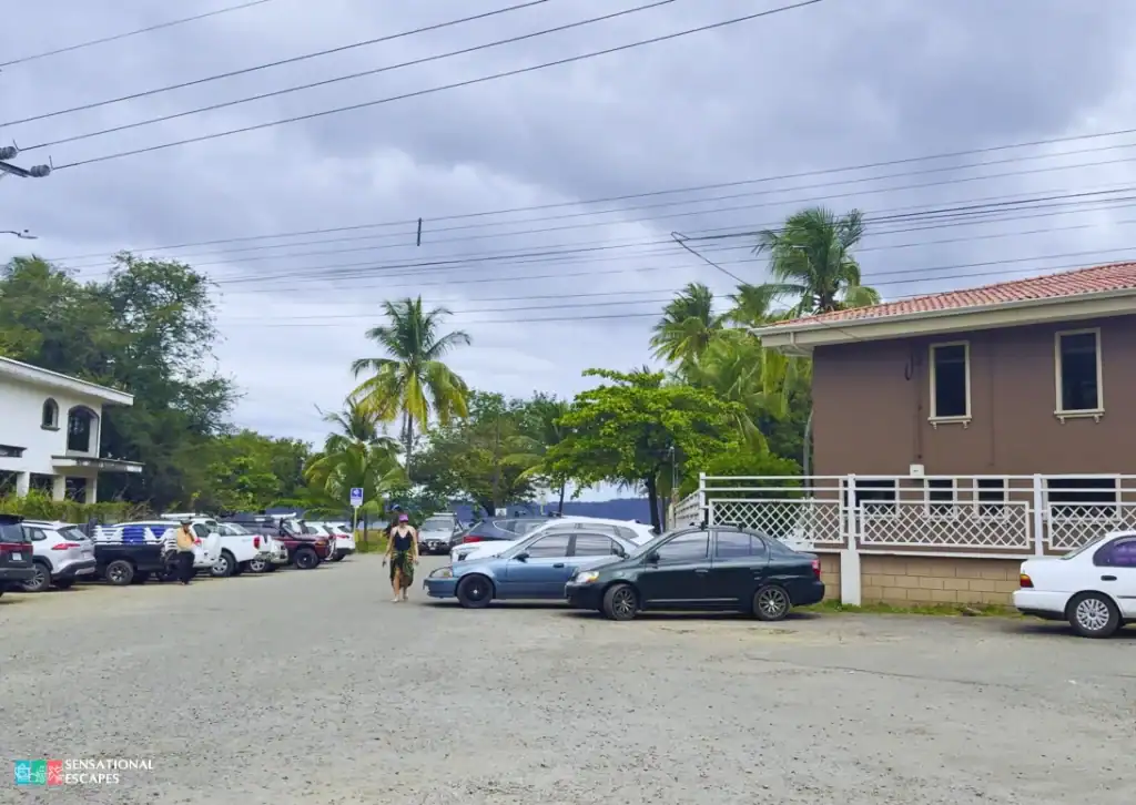 Acceso oeste a Playa Panamá en Guanacaste, cerca del restaurante Hacienda Blu, con un área de parqueo de lastre con autos, palmeras y vista hacia la entrada de la playa.