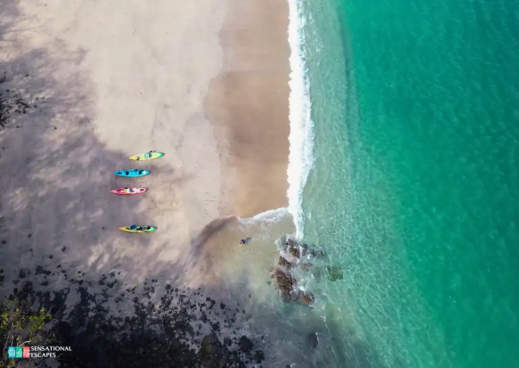Vista cenital de Playa Calz&oacute;n de Pobre con kayaks de colores alineados en la arena y olas turquesa suaves; una persona nada cerca de un peque&ntilde;o grupo de rocas.