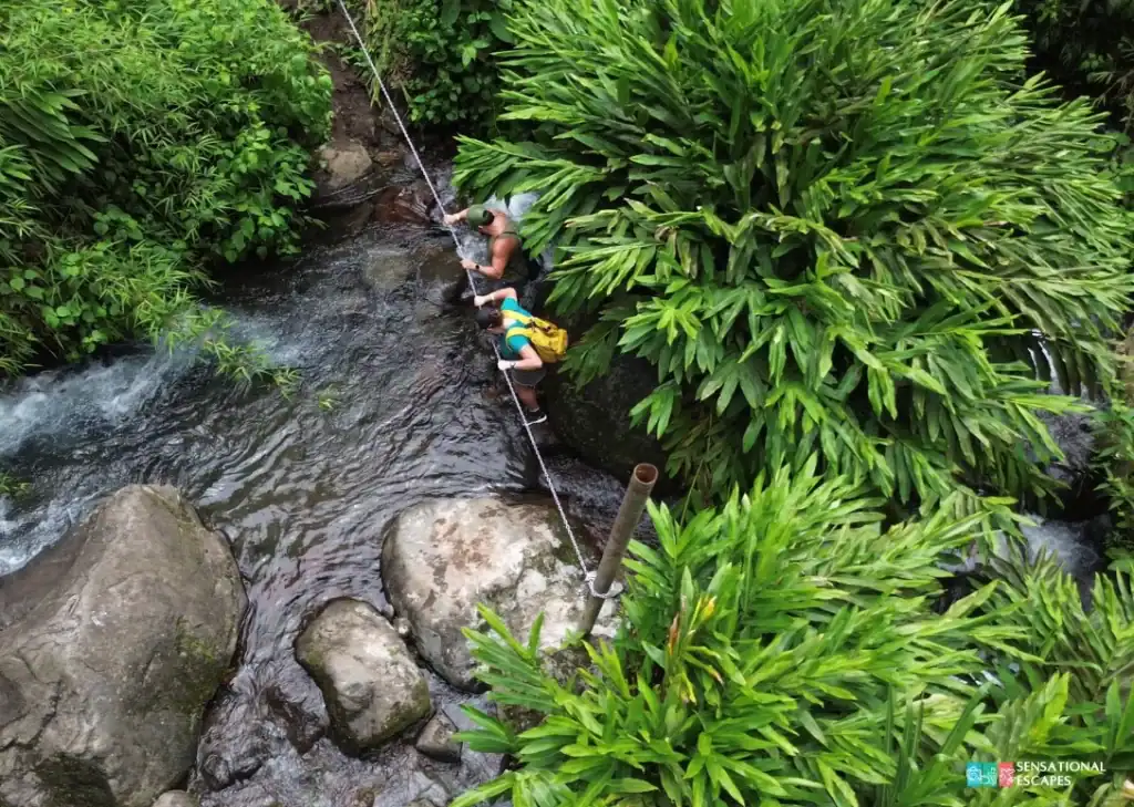 Two hikers using a rope to cross a shallow river surrounded by lush greenery on the trail to Las Trillizas Waterfalls in Turrialba, Costa Rica.