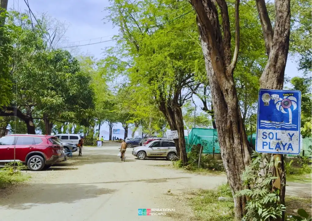 Entrada de camino de tierra a Playa Hermosa con carros parqueados, árboles con sombra, una persona en bicicleta y un rótulo azul gastado de “Sol y Playa” en un árbol.