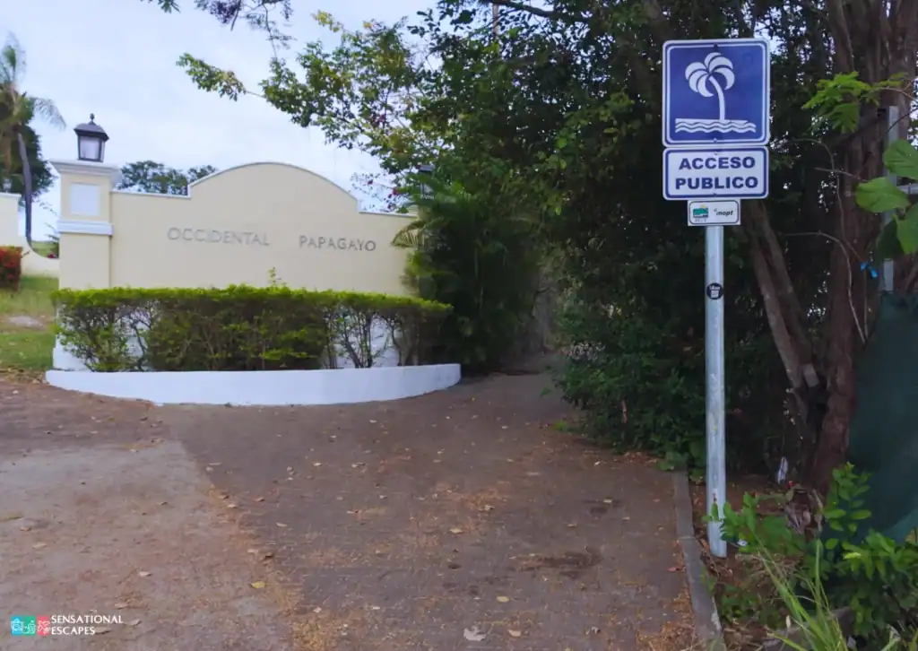R&oacute;tulo de acceso p&uacute;blico a la playa cerca de la entrada del Occidental Papagayo; sendero sombreado entre &aacute;rboles.