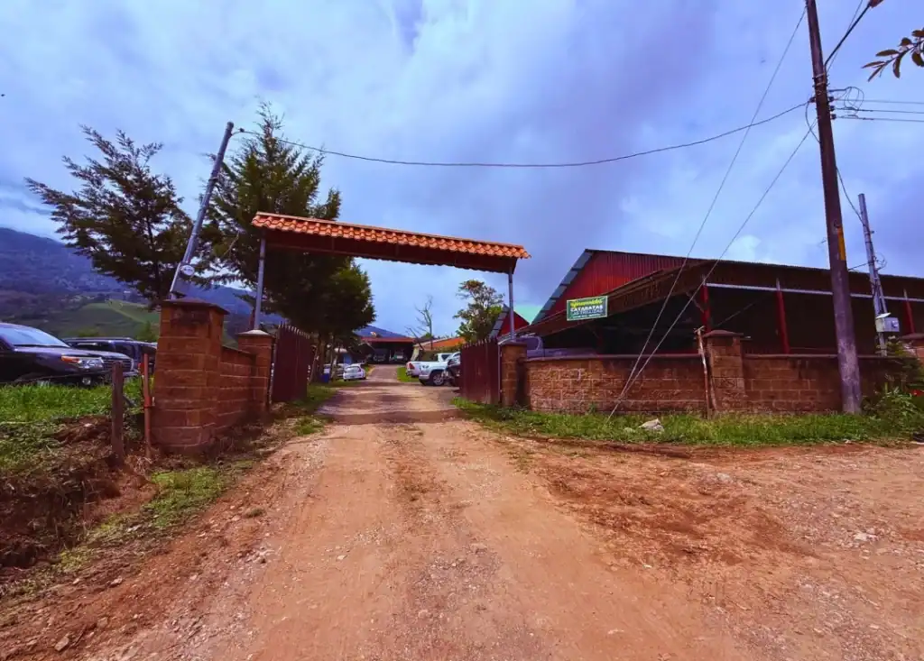 Gated entrance and reception area for Las Trillizas Waterfalls in Turrialba, Costa Rica, featuring a dirt road, parked cars, and a sign for the waterfalls next to a red-roofed building.