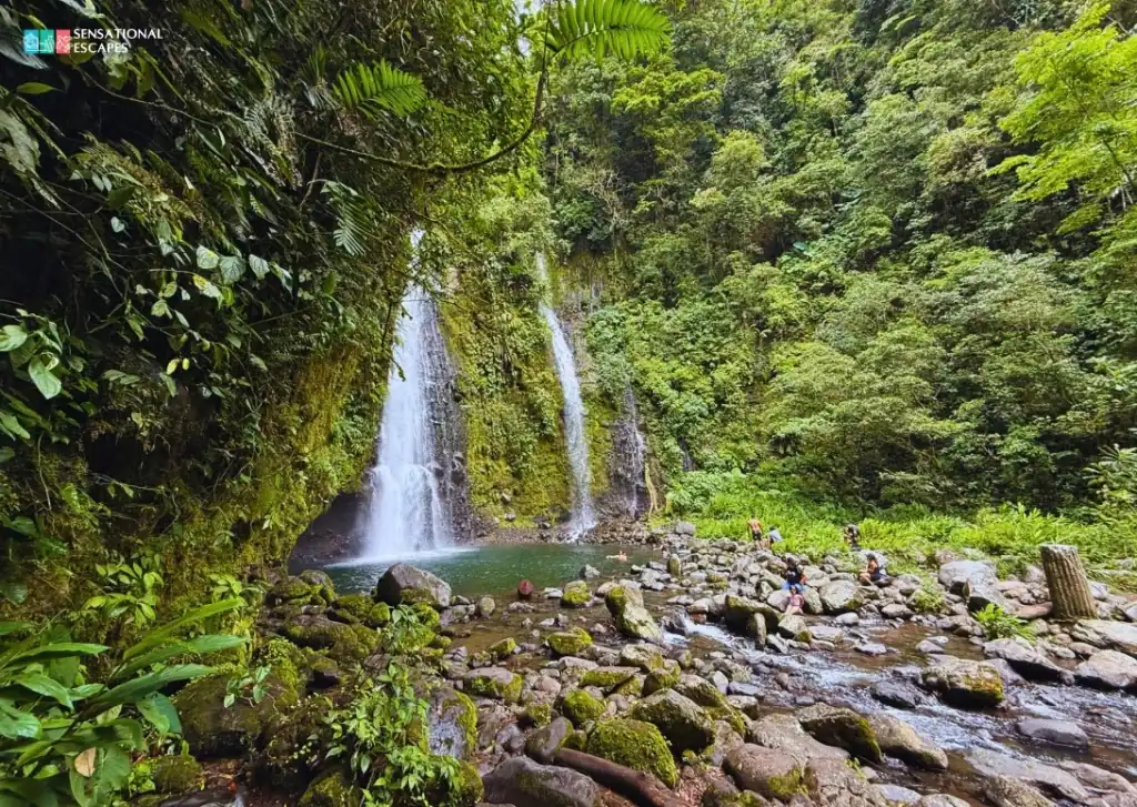 Twin waterfalls cascading into a rocky pool surrounded by dense jungle at Las Trillizas Waterfalls in Turrialba, Costa Rica, with hikers relaxing by the water's edge