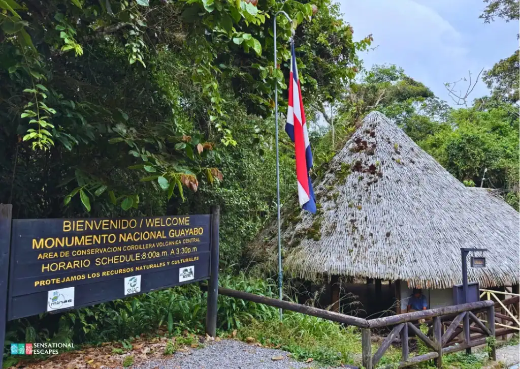 Entrance sign for Guayabo National Monument in Turrialba, Costa Rica, beside a thatched-roof hut and a tall flagpole flying the Costa Rican flag, surrounded by lush tropical greenery.