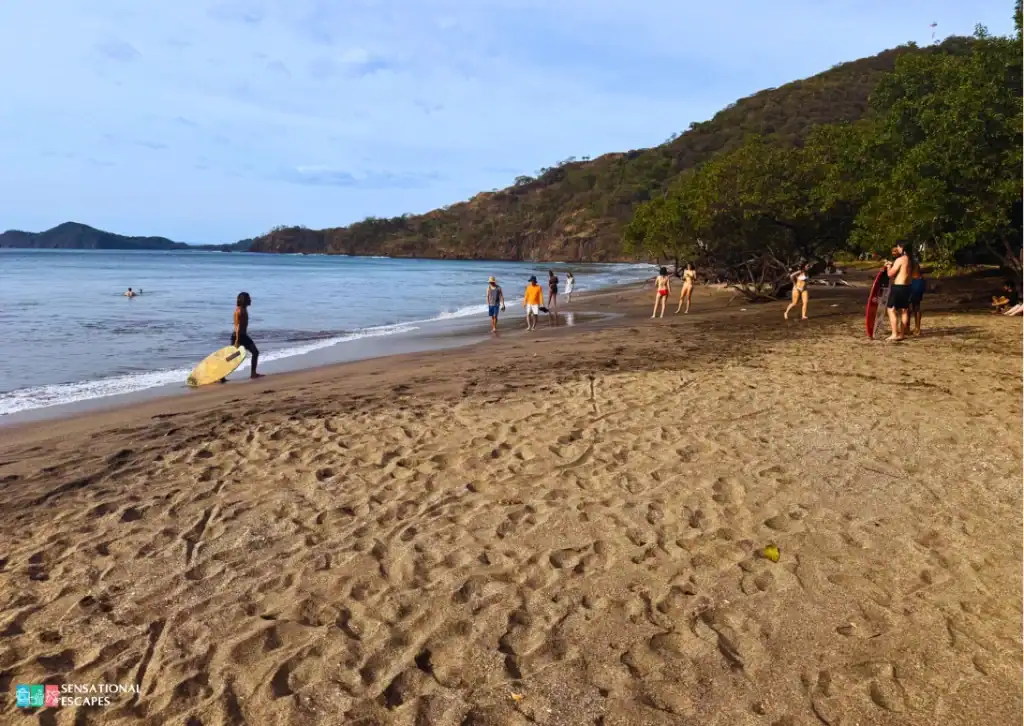 Gente disfrutando la orilla en Playa Hermosa: algunas personas caminan, otras con bodyboards o de pie cerca del agua; oleaje suave y cerros arbolados al fondo.