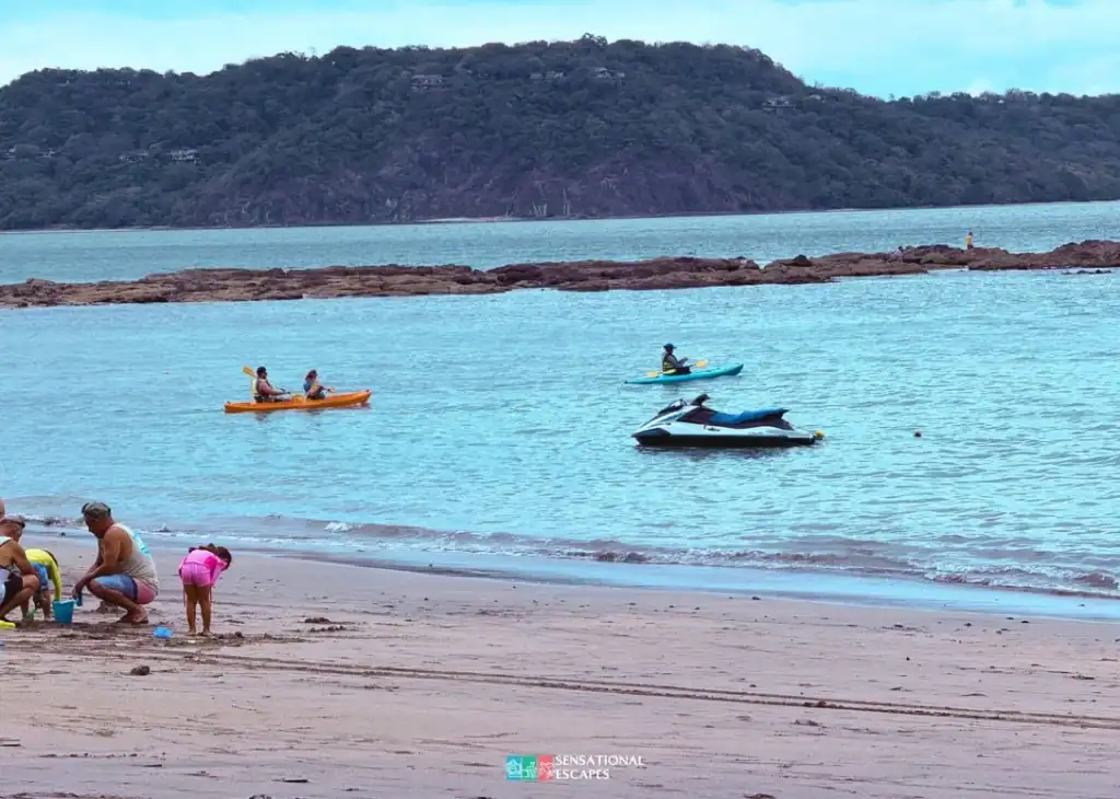 Gente en kayak y una moto acu&aacute;tica en las aguas calmas de Playa Buena; familias jugando en la arena.
