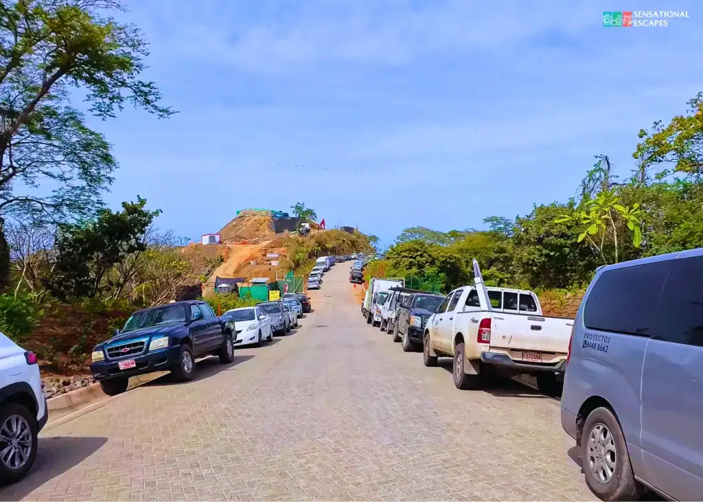 Carretera principal hacia Playa Penca en Guanacaste, con carros estacionados a la orilla, subiendo entre vegetación y zona en construcción bajo cielo azul.