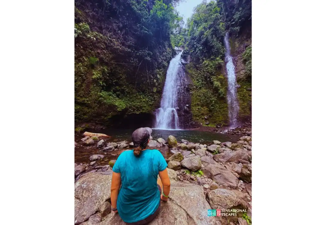 Visitor sitting on rocks and admiring the twin cascades of Las Trillizas Waterfalls in Turrialba, Costa Rica, surrounded by lush greenery and a natural pool.
