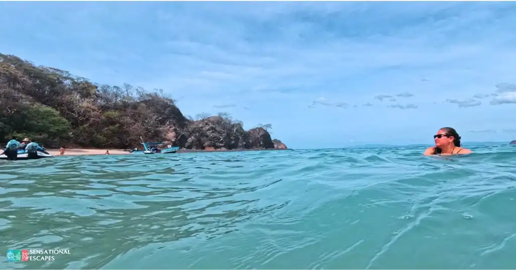 Persona disfrutando del agua turquesa y clara en Playa Penca, con acantilados rocosos, botes y algunas personas cerca de la costa boscosa.