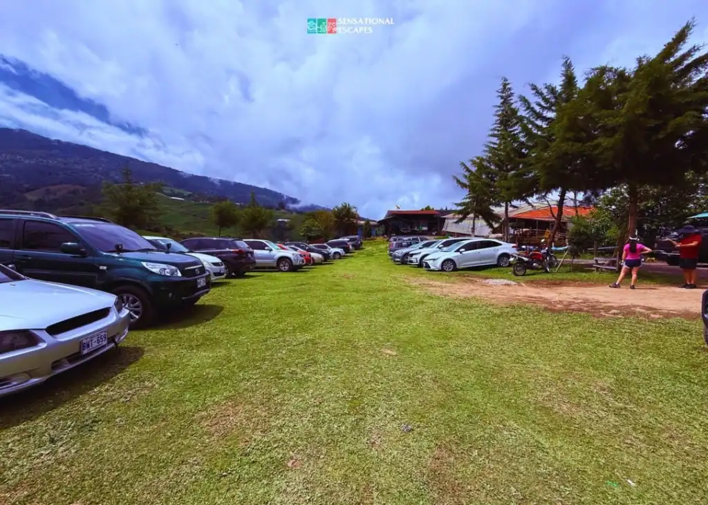 Grassy parking lot at Las Trillizas Waterfalls in Turrialba, Costa Rica, filled with cars and surrounded by mountains and trees, near the reception area.