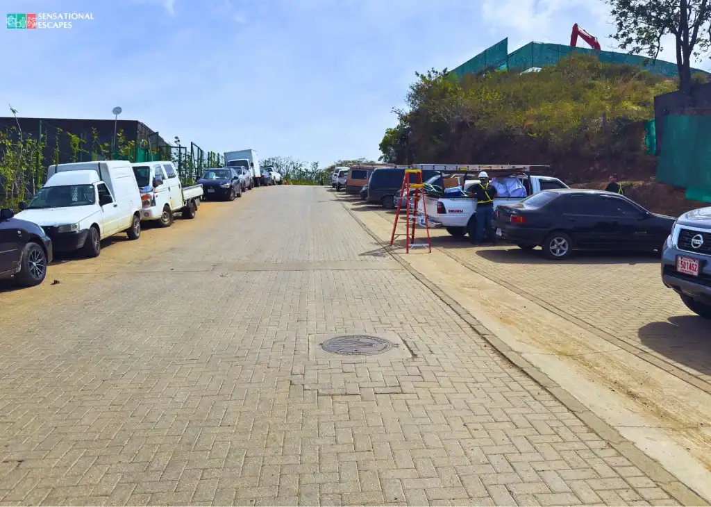 Área de parqueo para Playa Penca en Guanacaste con carros y camiones de obra a la par de una calle asfaltada junto a una zona en construcción.