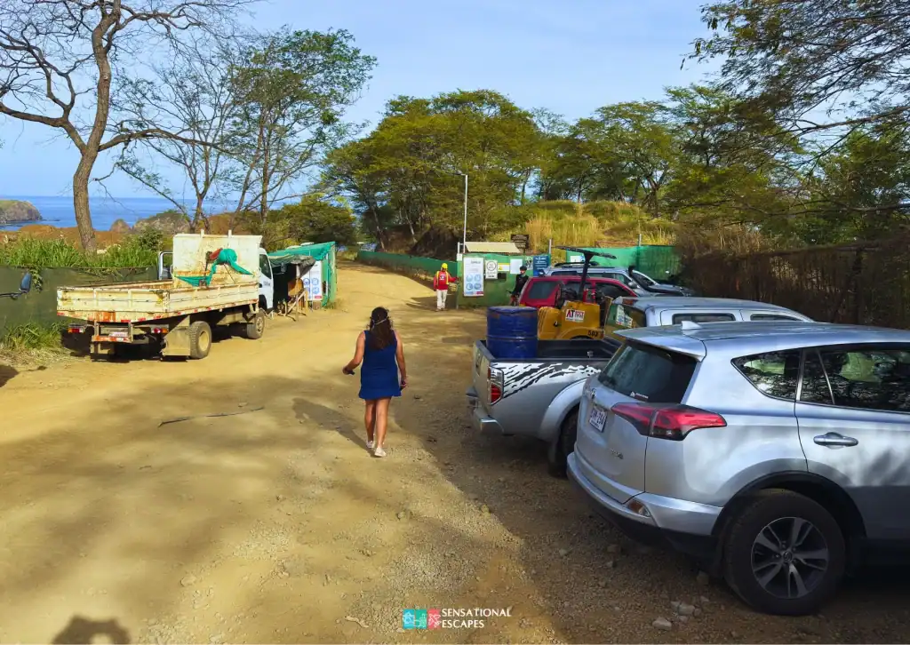 &Aacute;rea de parqueo de tierra cerca de la entrada al sendero de Playa Calz&oacute;n de Pobre, con varios carros, algunos trabajadores y una mujer con vestido azul caminando hacia la playa.