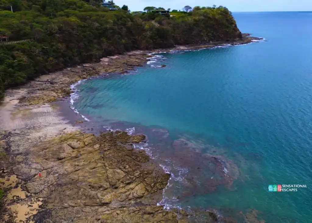 Extremo oeste rocoso de Playa Buena con agua turquesa y pozas de marea; vegetaci&oacute;n verde a lo largo de la costa.