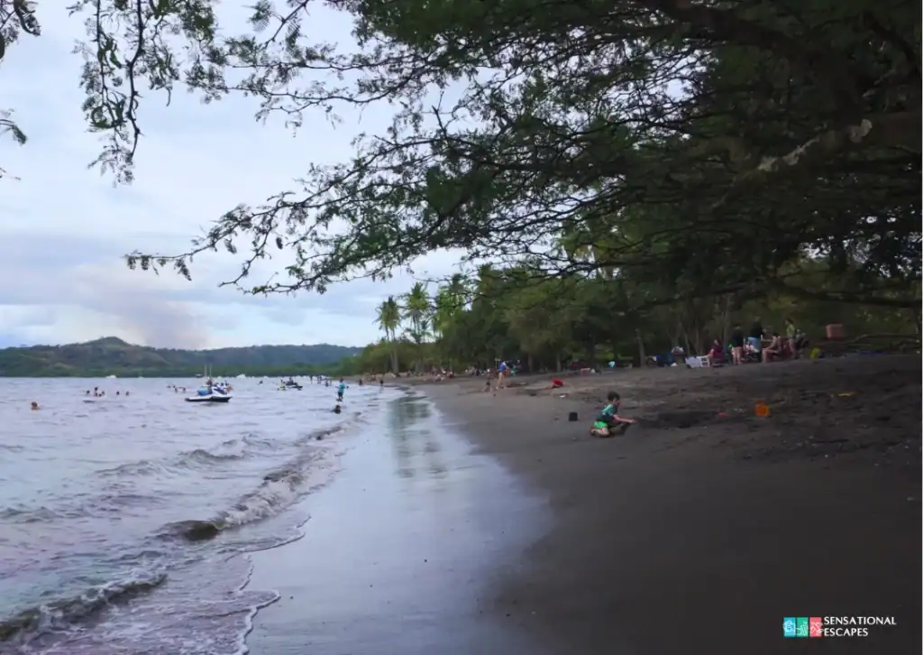 Mucha gente disfruta la orilla y las aguas tibias en Playa Panamá, Guanacaste; niños juegan en la arena y se ven jet skis a lo lejos bajo una playa arbolada.