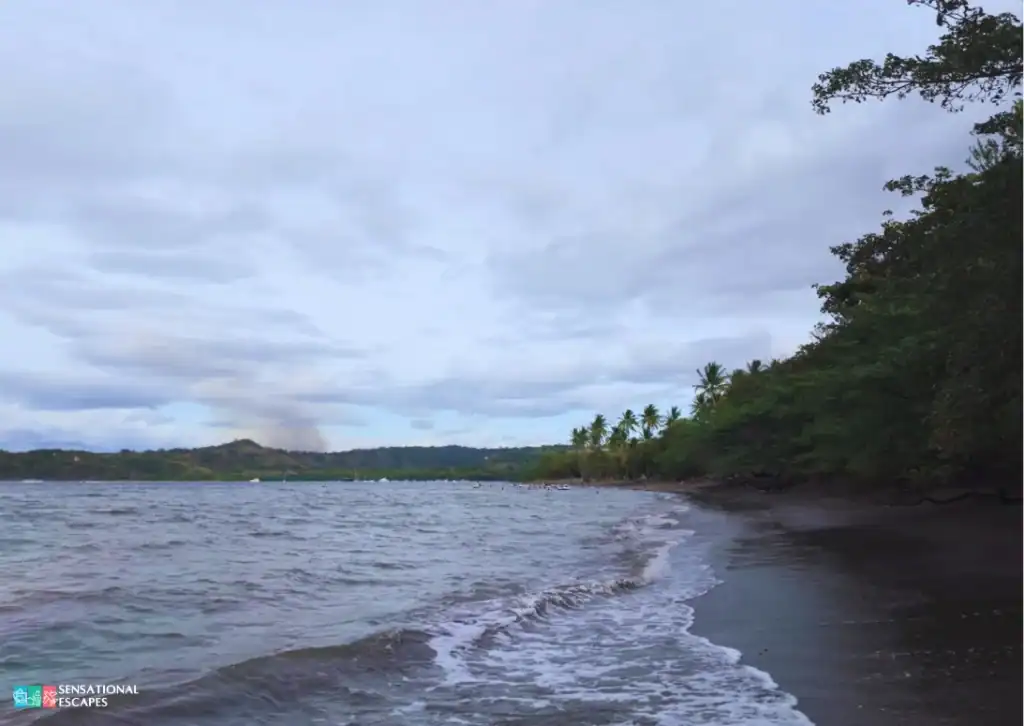 Vista del lado oeste de Playa Panamá en Guanacaste, Costa Rica, con olas suaves, arena oscura, vegetación frondosa y una vista lejana del Golfo de Papagayo bajo un cielo nublado.