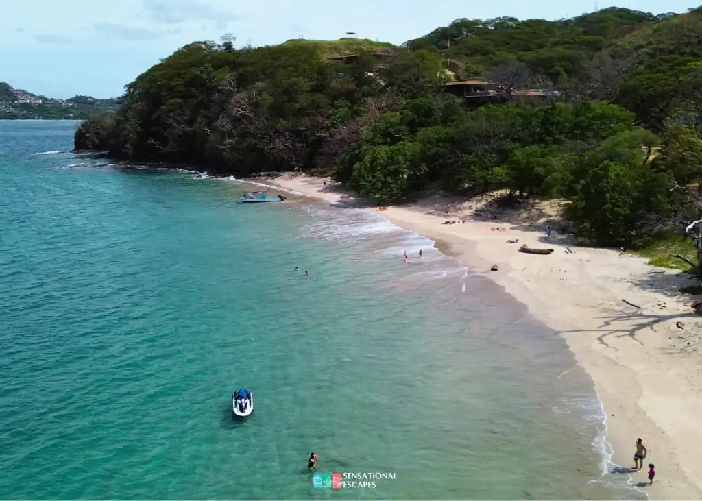 Vista aérea de Playa Penca en Guanacaste con agua turquesa clara, arena dorada, algunos visitantes y botes cerca de la orilla, rodeada de vegetación.