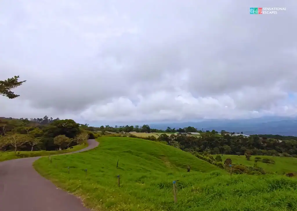 Winding rural road through lush green hills leading toward Guayabo National Monument in Turrialba, Costa Rica, under a cloudy sky with distant mountain views.