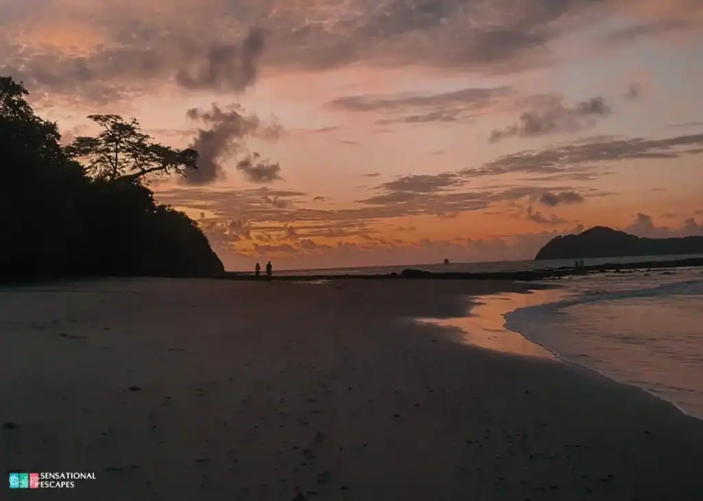 Atardecer sereno en Playa Buena con cielo naranja y rosado reflejado en la arena mojada; siluetas caminando por la orilla.