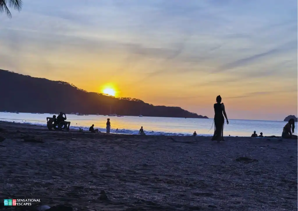 Siluetas de personas descansando en la playa durante un atardecer dorado en Playa Hermosa, con olas suaves y botes anclados al fondo.