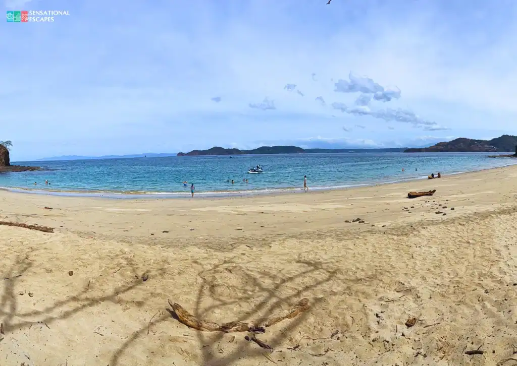 Amplia orilla arenosa en Playa Penca con agua turquesa calma, gente nadando y caminando, y una moto acuática frente a la costa con el Golfo de Papagayo a lo lejos.