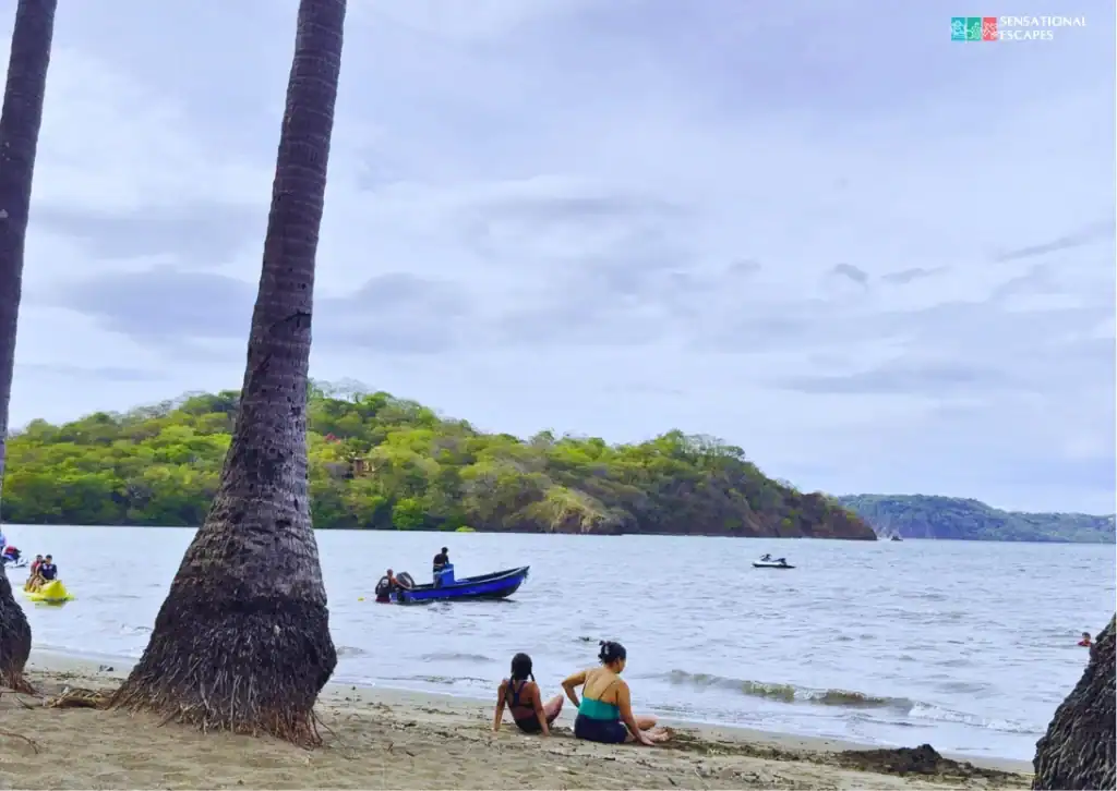 Personas descansan bajo palmeras en Playa Panamá, Guanacaste; hay botes y jet skis en el agua tranquila y una ladera verde al fondo.